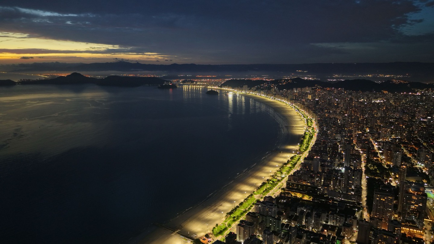 Vista aérea noturna da orla de Santos (SP), com a praia e o jardim da orla iluminados ao longo da baía e prédios residenciais à direita