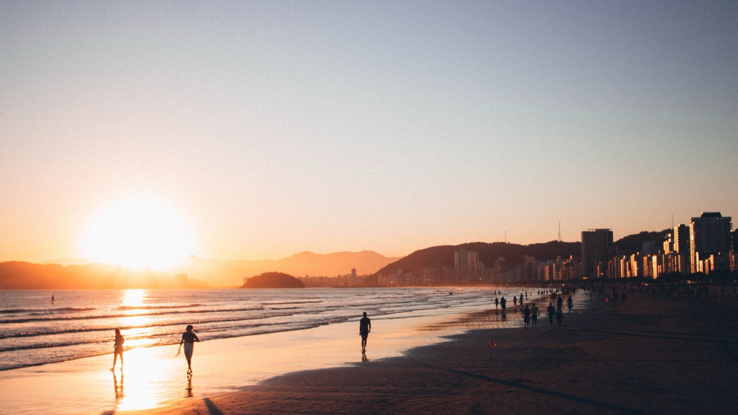 Pôr do sol na praia do Boqueirão, em Santos, com pessoas caminhando na areia molhada e os prédios da orla iluminados pela luz dourada