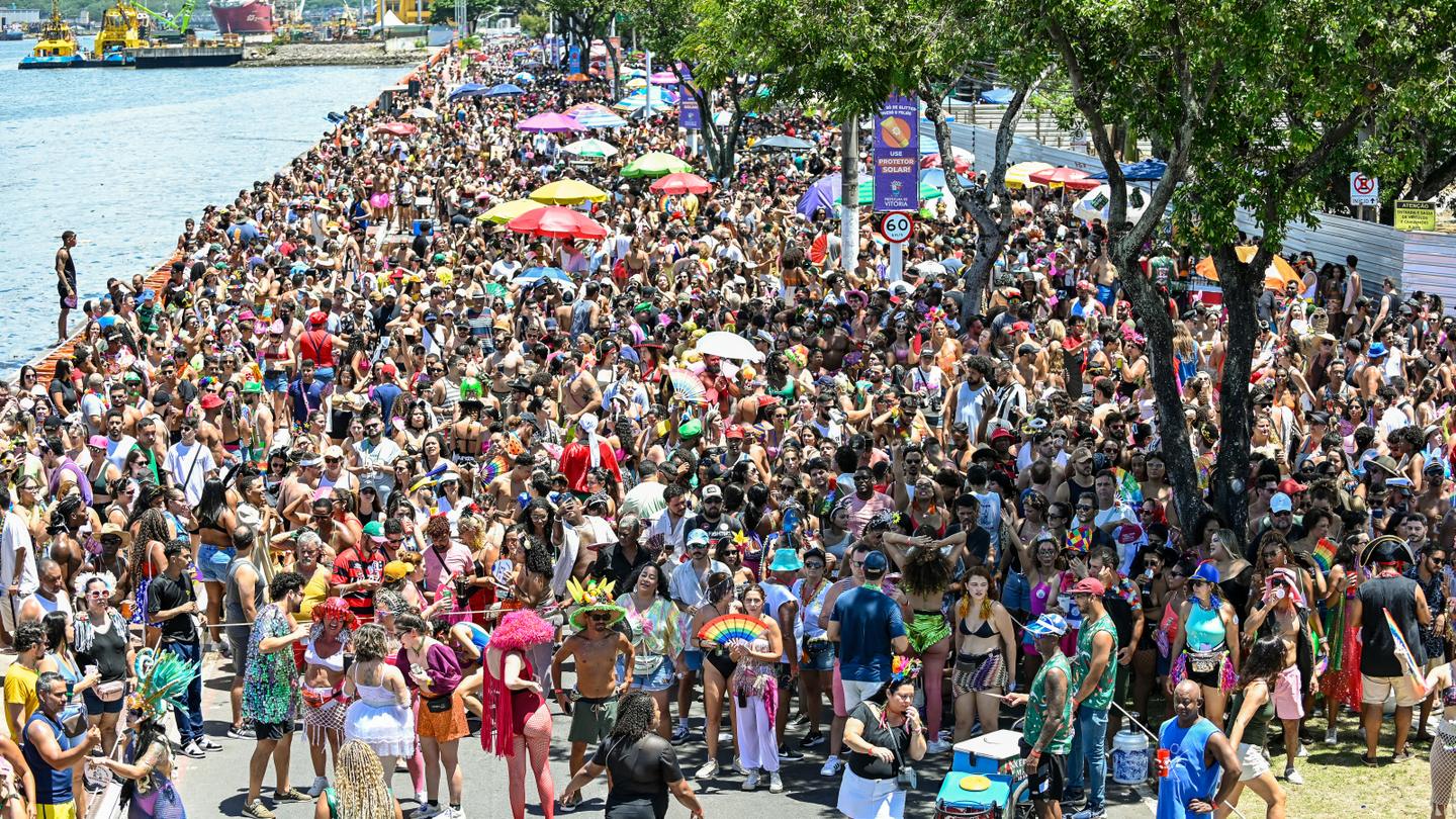 Multidão de foliões no bloco Regional da Nair, na Avenida Beira-Mar, no Centro de Vitória, com a baía e o porto ao fundo, num dia de sol no Carnaval de 2025.