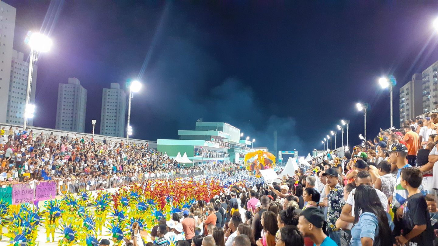 Desfile da escola de samba Unidos dos Morros na Passarela do Samba Dráuzio da Cruz, em Santos (SP), com alas coloridas, arquibancadas lotadas e refletores acesos à noite.