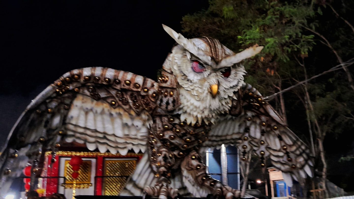 Carro abre-alas da escola de samba Unidos de Jucutuquara, com uma coruja gigante de asas abertas, na concentração do Sambão do Povo, em Vitória (ES), à noite.