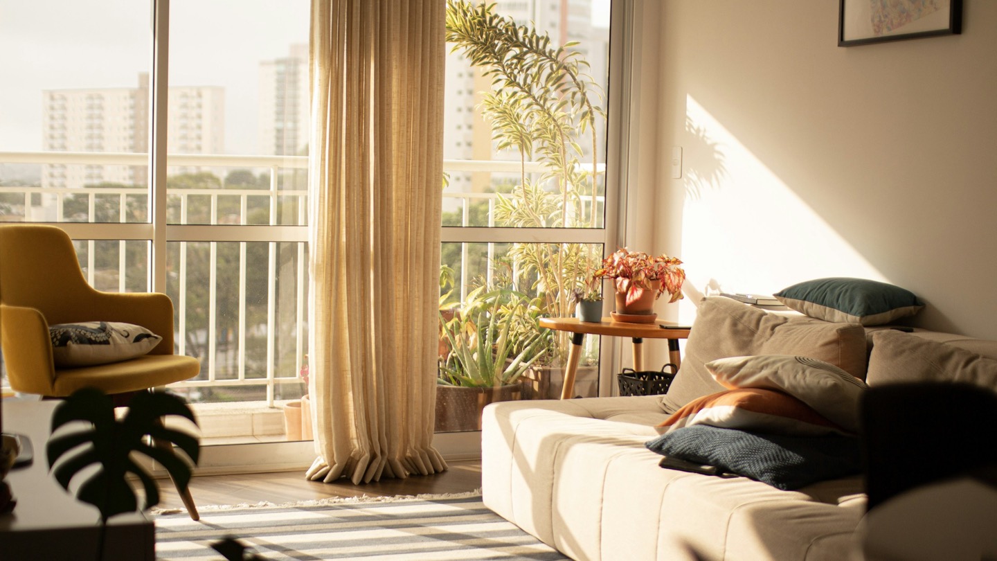 Sala de estar banhada pelo sol do fim de tarde, com sofá bege, poltrona mostarda, cortina de linho e uma varanda cheia de plantas com prédios residenciais ao fundo