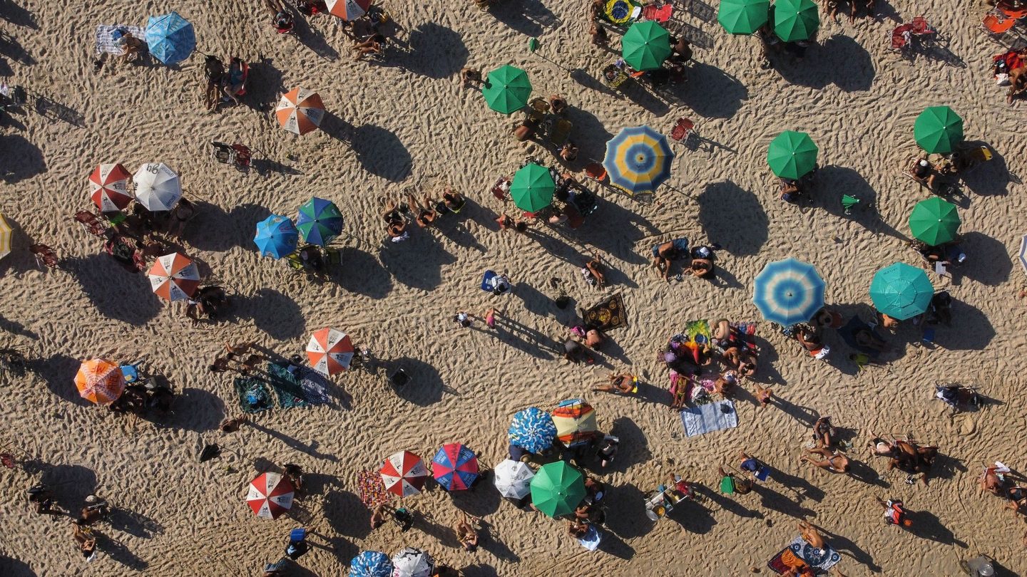 Praia lotada em dia de sol vista de cima: guarda-sóis coloridos, cangas e cadeiras na areia, banhistas pequenos e sombras longas de fim de tarde.