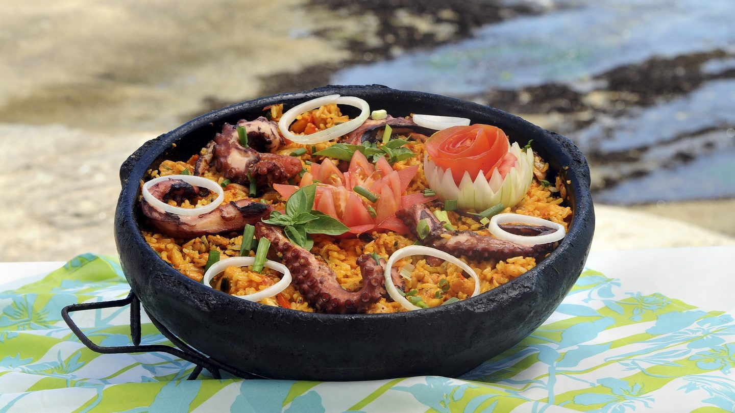 Arroz de polvo servido em panela de barro preta sobre toalha estampada, em mesa à beira-mar, com areia, pedras e mar desfocados ao fundo.