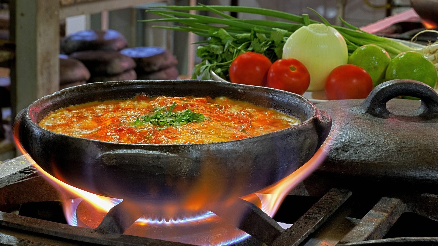 Moqueca capixaba fervendo em panela de barro preta sobre a chama do fogão, com tomates, cebola, limões e cheiro-verde ao fundo.