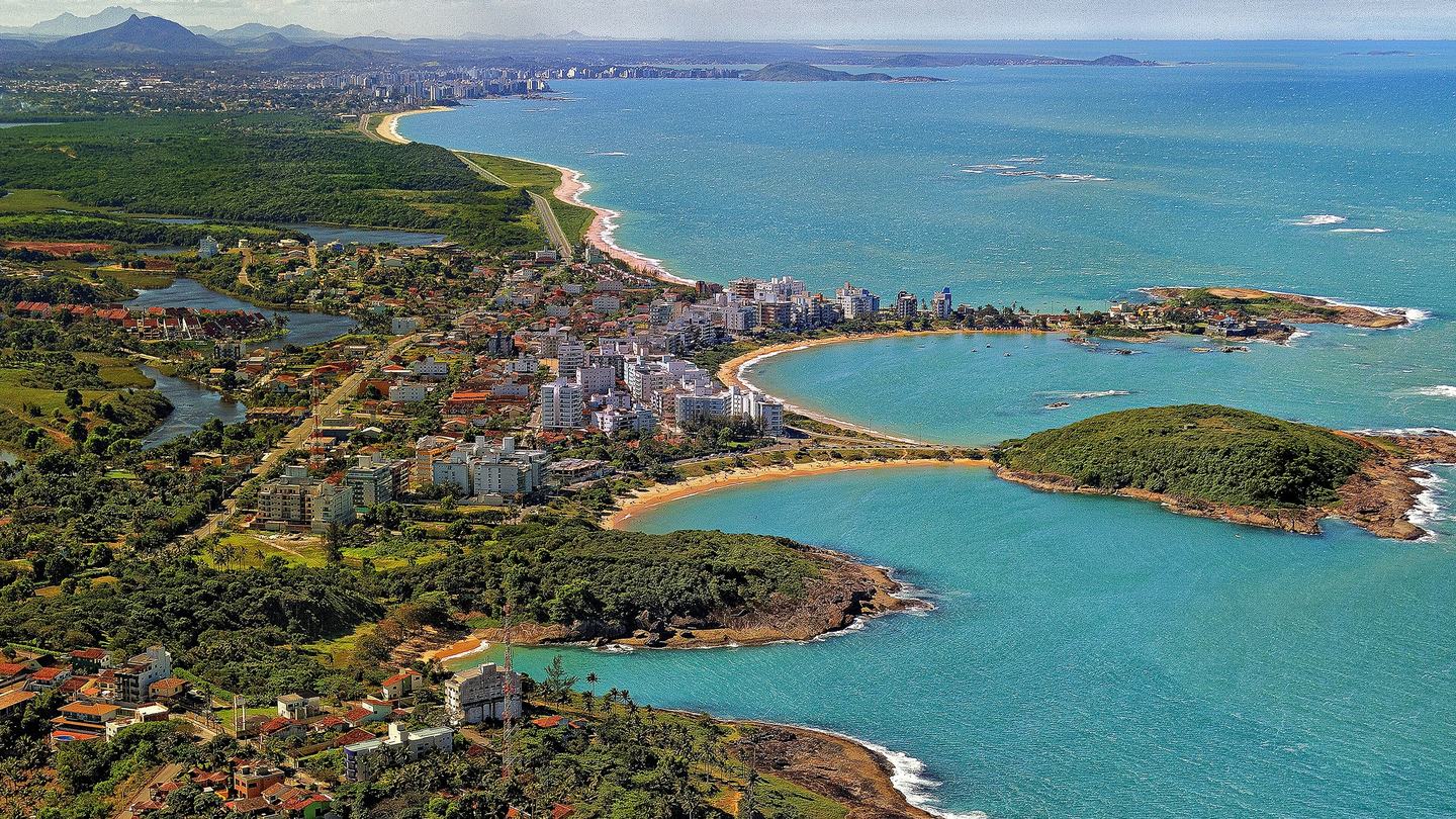 Vista aérea das enseadas de águas turquesa da Enseada Azul, em Guarapari (ES), com ilhota coberta de mata, morros verdes e a orla da cidade ao fundo.