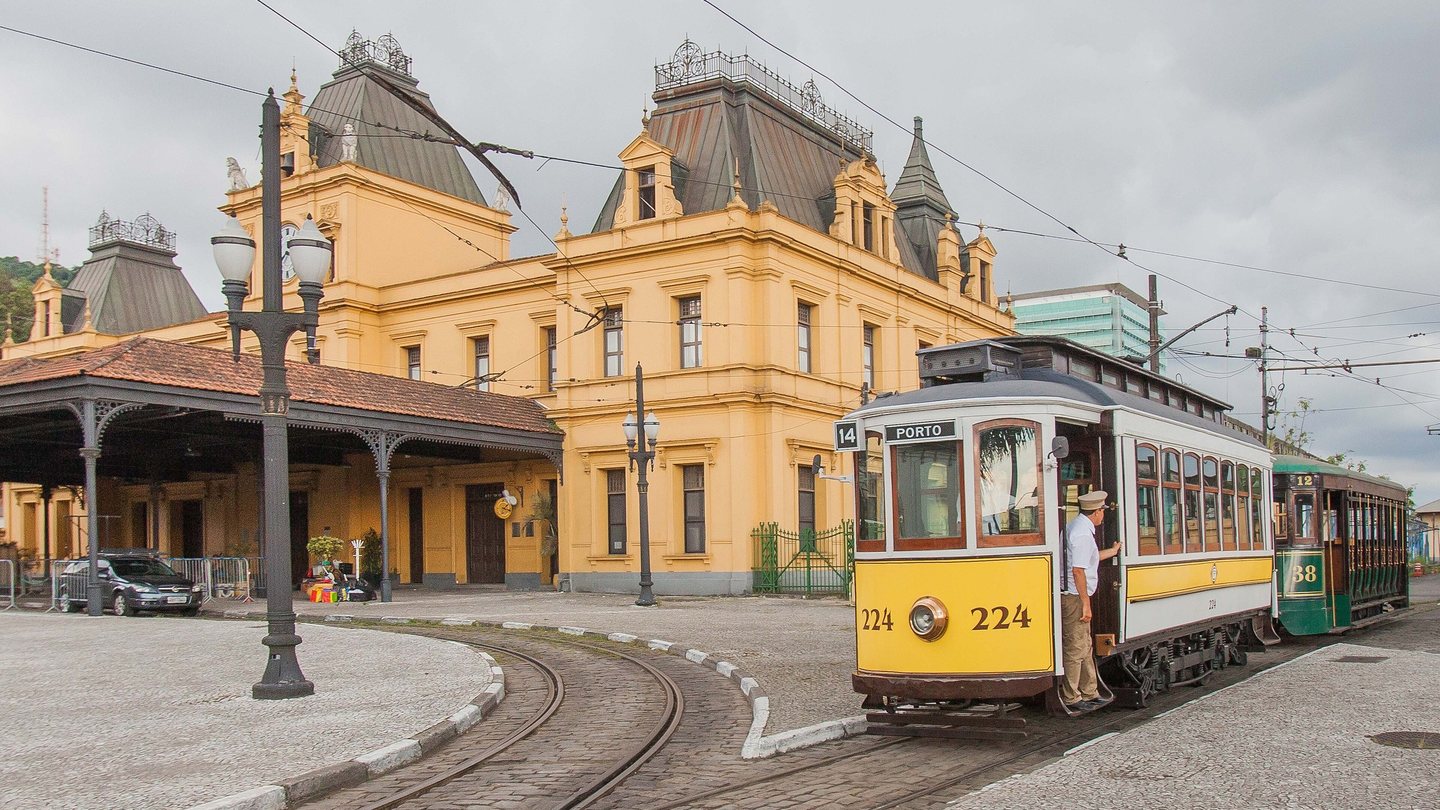 Bonde turístico amarelo e branco nos trilhos em frente à Estação do Valongo, prédio eclético amarelo no centro histórico de Santos (SP).