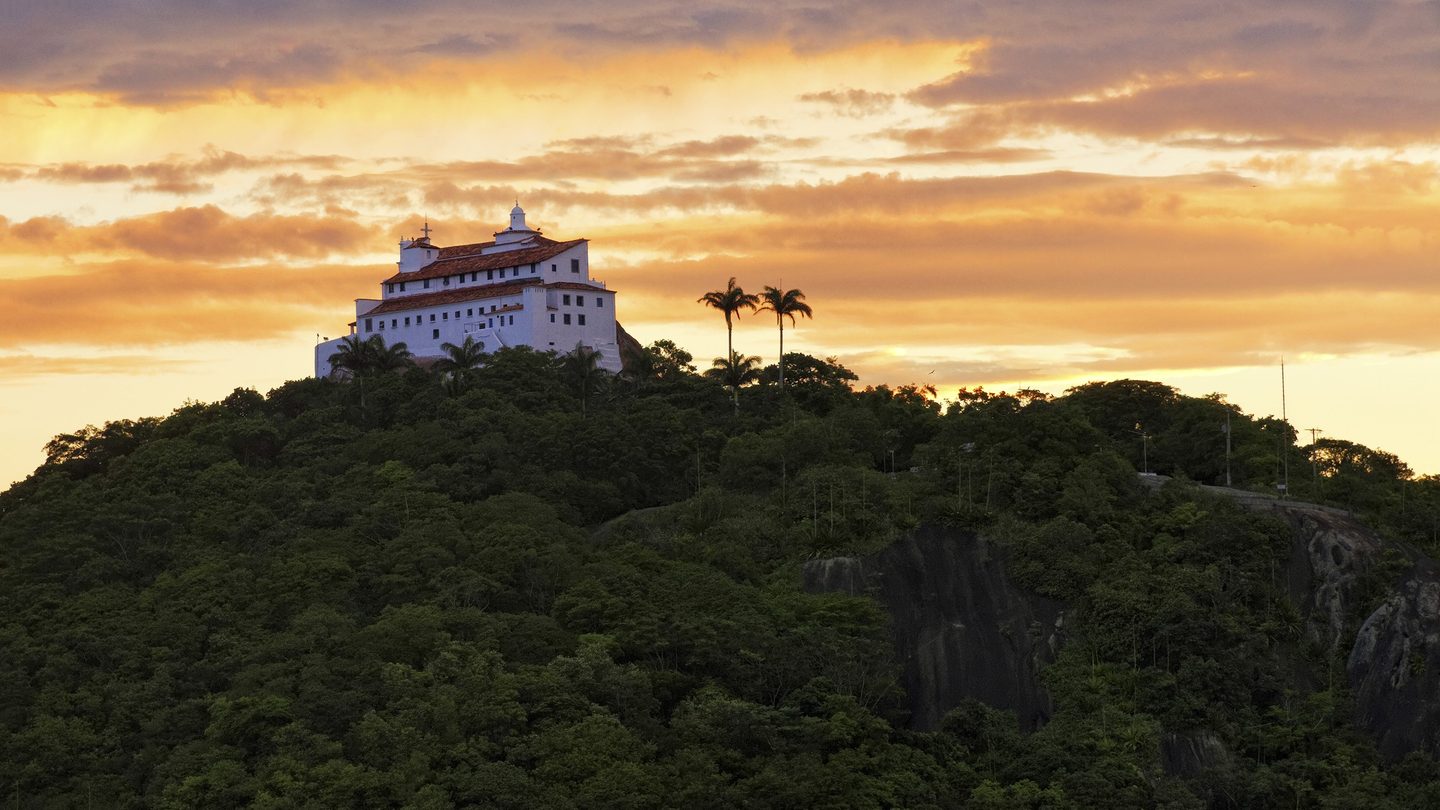 Convento da Penha no alto do morro coberto de Mata Atlântica, em Vila Velha (ES), sob um céu dourado de pôr do sol, com duas palmeiras-imperiais ao lado.