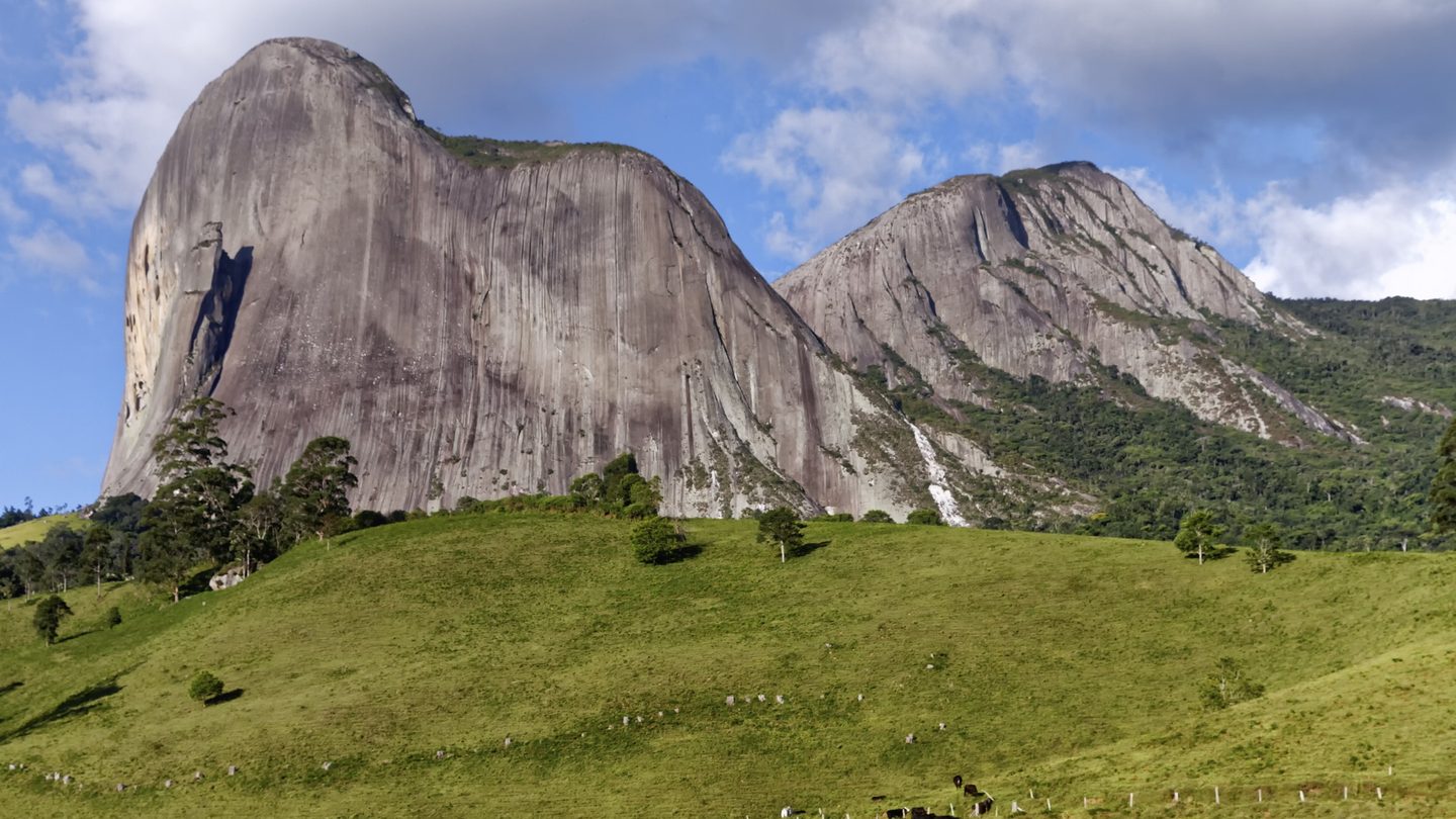 A Pedra Azul e os paredões vizinhos no Parque Estadual da Pedra Azul, em Domingos Martins (ES), erguendo-se sobre pastos verdes com gado sob céu azul com nuvens.