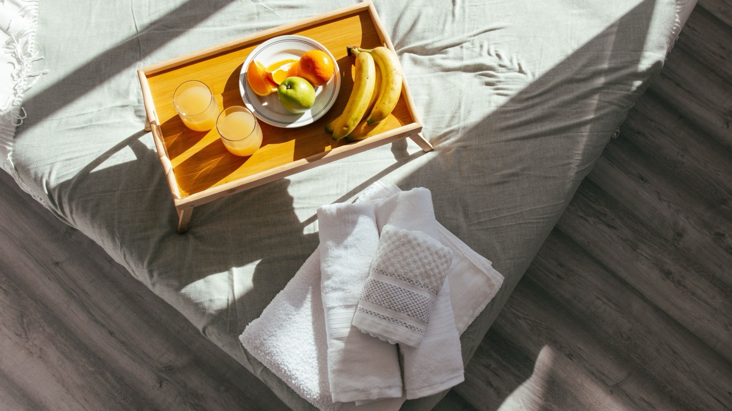 Vista de cima de uma cama com manta verde-acinzentada, uma bandeja de madeira com dois copos de suco, frutas e bananas, e uma pilha de toalhas brancas dobradas, iluminadas pela luz do sol que entra pela janela.