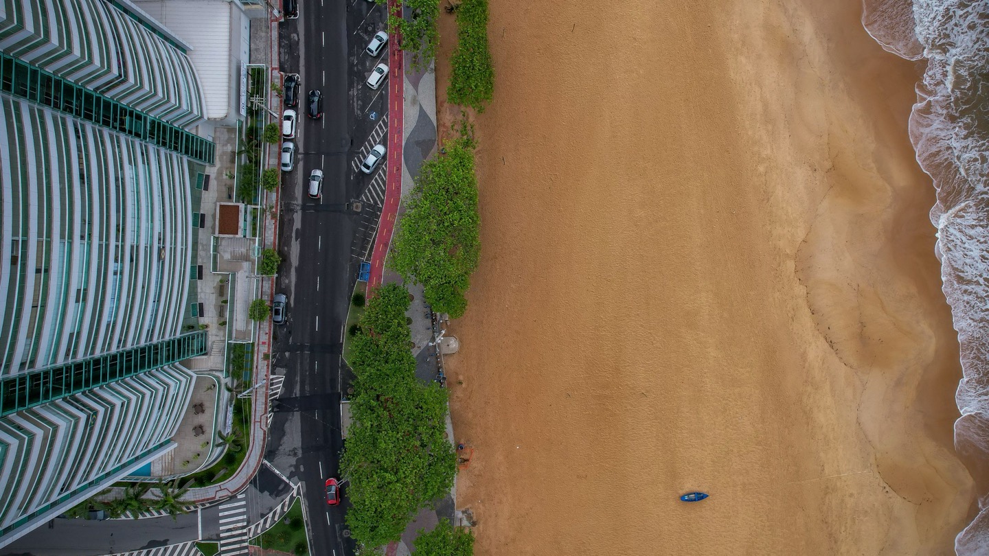Vista aérea vertical da orla de Vila Velha (ES), com prédio residencial à beira-mar, avenida, ciclovia, calçadão arborizado e faixa de areia até o mar