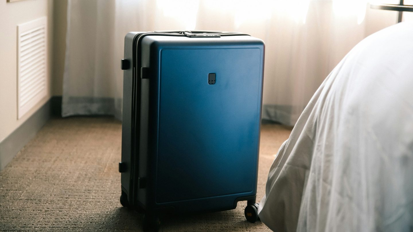 Mala de rodinhas azul em pé no carpete de um quarto de hotel, ao lado da cama com lençol branco, diante de uma cortina translúcida iluminada pela luz do dia.