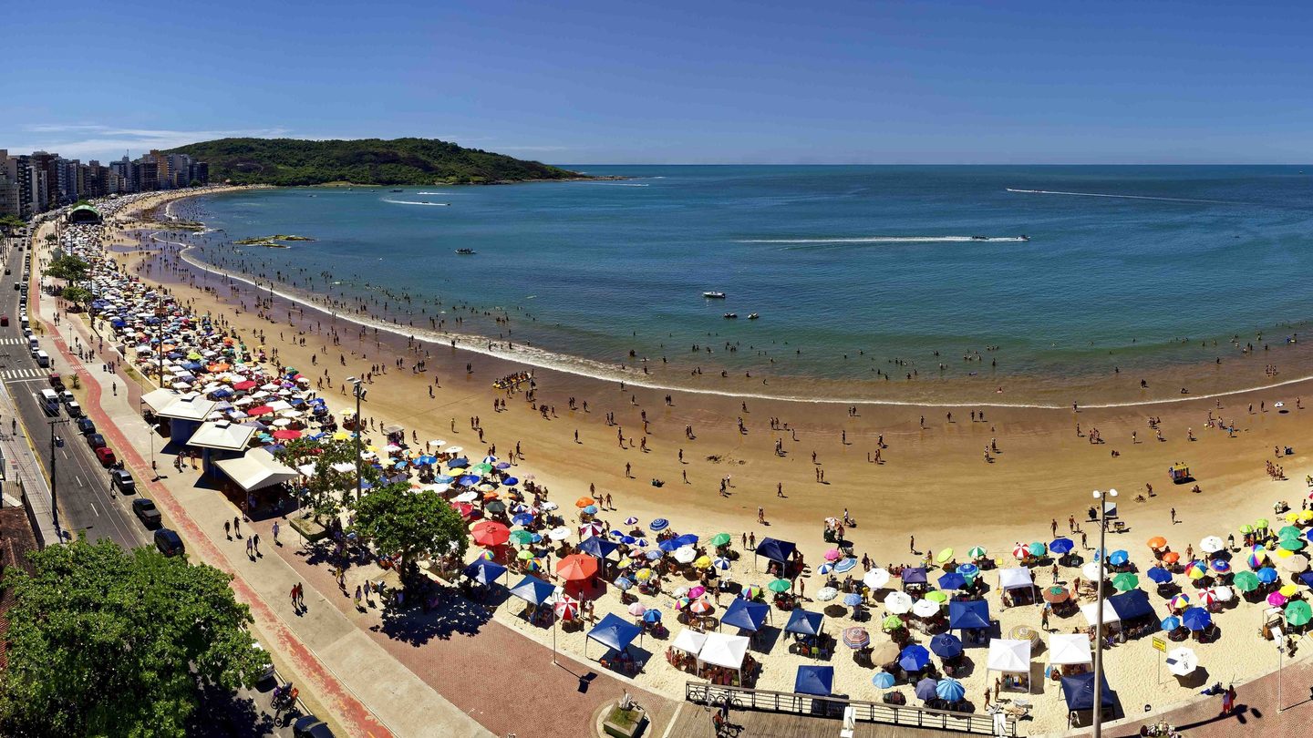 Praia do Morro, em Guarapari (ES), lotada em dia de verão, vista do alto: guarda-sóis coloridos e tendas na areia, banhistas no mar, calçadão e avenida à beira-mar, prédios e o Morro da Pescaria ao fundo.
