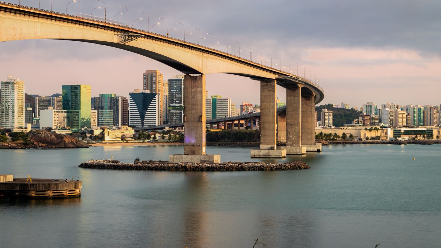 Terceira Ponte em curva sobre a baía de Vitória ao amanhecer, com céu em tons de rosa e lilás, água calma e os prédios da Enseada do Suá ao fundo, em Vitória (ES).